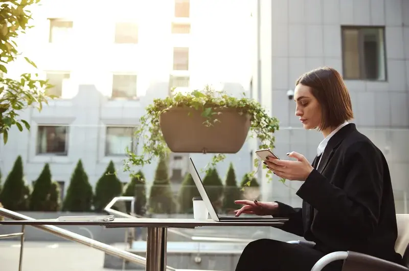 Mujer trabajando al aire libre usando una laptop y un teléfono móvil, mejorando su productividad laboral